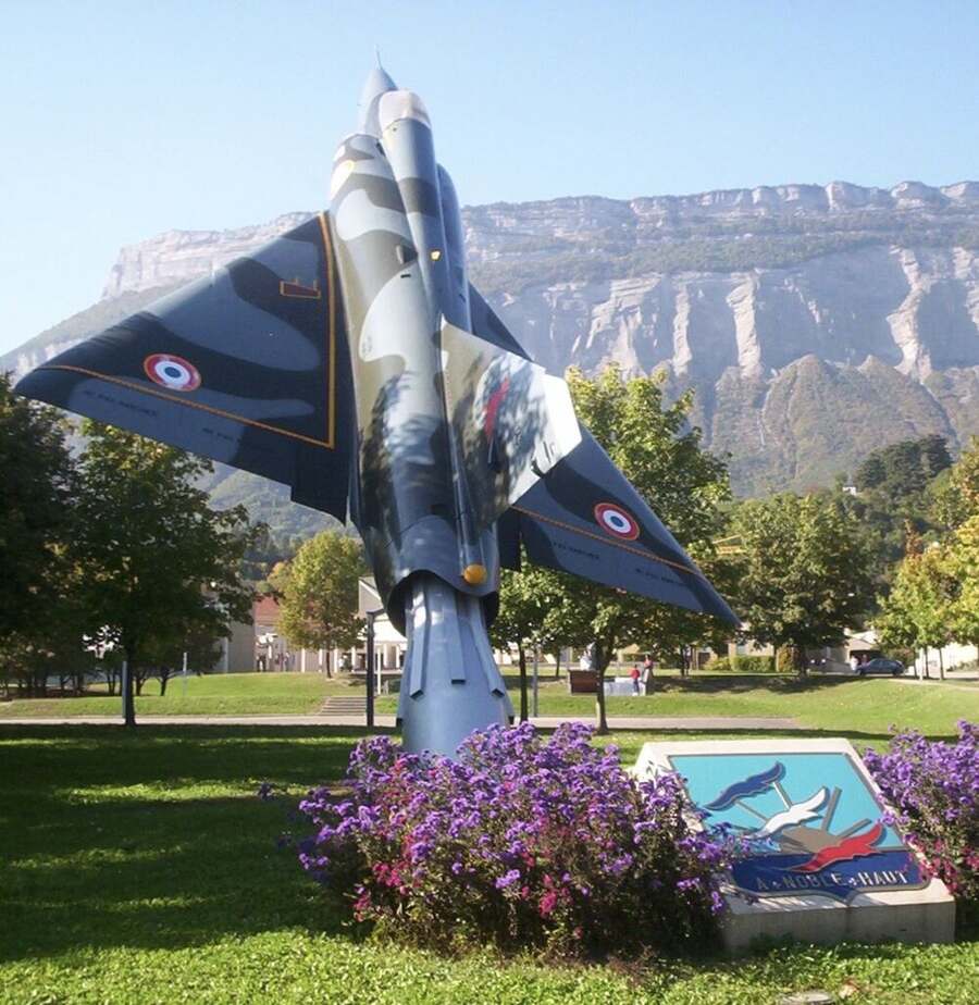 Mirage III monté en monument devant un massif montagneux, insigne de l'école au premier plan